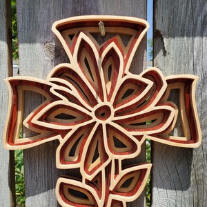 May include: A layered wooden cross with a floral design. The cross is made of alternating layers of light and dark wood, creating a three-dimensional effect. The floral design is centered on the cross, with a stem and leaves at the bottom. The cross is hanging on a weathered wooden fence.
