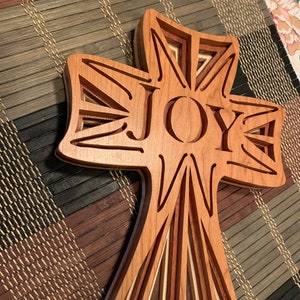 May include: A wooden cross with a layered design and the word "JOY" carved into the center. The cross is made of different shades of wood, creating a multi-dimensional effect.