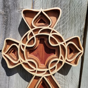 May include: A layered wooden cross with a detailed design. The cross features a combination of light and dark wood tones, with intricate cutouts and a multi-dimensional effect. The cross is displayed against a weathered wooden background.