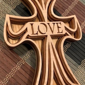 May include: A wooden cross with a layered design, featuring the word "LOVE" carved into the center. The cross is made of light brown wood and has a natural finish.