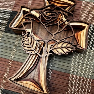 Peut inclure: Une croix en bois avec une rose sculptée au centre. La croix est faite de plusieurs couches de bois, créant un effet 3D. La rose est détaillée et complexe, avec des feuilles et des pétales.