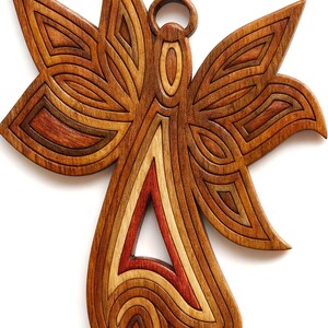May include: Wooden angel wall art with layered design. The angel features a detailed wing and body design with a mix of light and dark wood tones, including red and brown. The angel has a circular loop at the top for hanging.