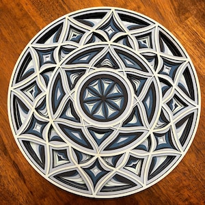May include: A white and blue paper mandala with intricate geometric patterns. The mandala is layered and has a 3D effect.