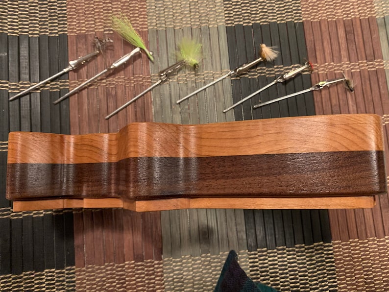 Puede incluir: Una caja de almacenamiento de madera para se&ntilde;uelos de pesca con mosca con un patr&oacute;n de veta de madera marr&oacute;n oscuro y marr&oacute;n claro. Seis se&ntilde;uelos de pesca con mosca se muestran en la parte superior de la caja.
