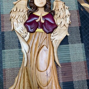 May include: Wooden angel sculpture with outstretched wings, carved from various wood tones. The angel has a serene expression, with hands clasped in prayer. The angel's dress is a light brown, the wings are a lighter wood, and the bodice is a deep burgundy.