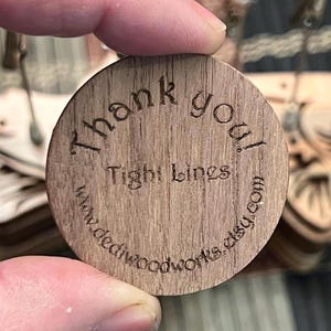 Puede incluir: Una etiqueta redonda de madera con el texto "&iexcl;Gracias!" y "Tight Lines" grabado en ella. La etiqueta tambi&eacute;n incluye la direcci&oacute;n del sitio web "www.deadwoodworks.etsy.com".