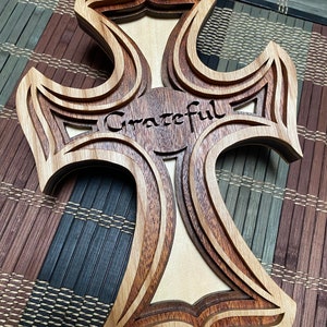Peut inclure: Une croix en bois avec un design en couches, comportant le mot "Grateful" gravé au centre. La croix est faite de bois clair et foncé, créant un effet contrasté.