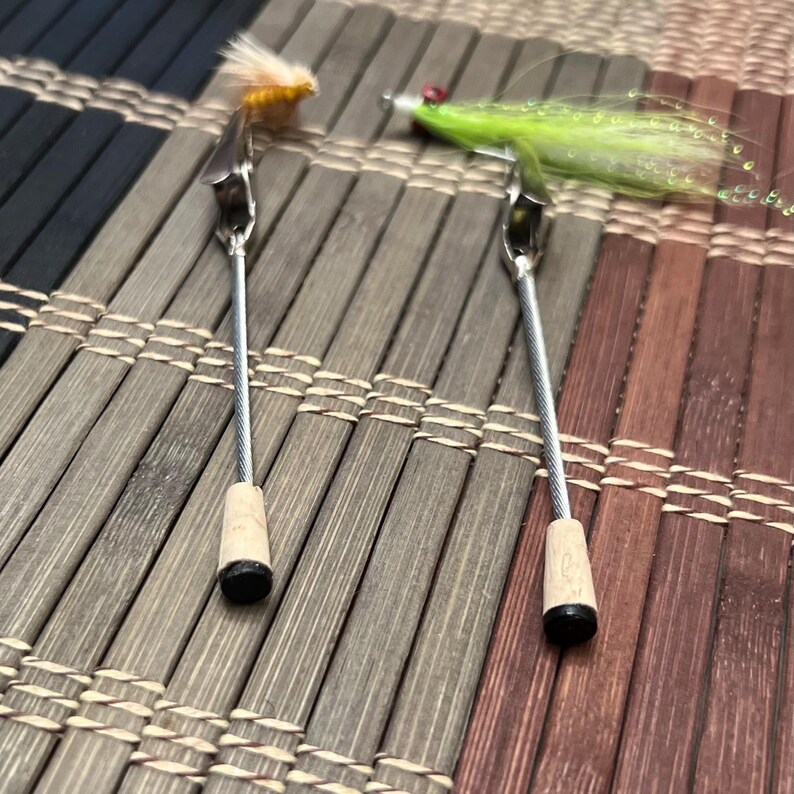 Puede incluir: Dos herramientas de atado de moscas de pesca con mosca con mangos de madera y clips de metal. Las herramientas sostienen dos moscas de diferentes colores.