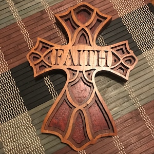May include: A layered wooden cross featuring the word "FAITH". The cross is crafted from various shades of brown and reddish-brown wood, with detailed decorative elements. It rests on a multi-coloured bamboo mat, showcasing the craftsmanship.