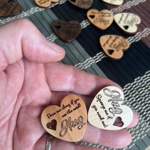 May include: Two wooden heart-shaped ornaments with the text "Pass me along if you see the need! Hug" and "Squeeze me if you need me! Hug" engraved on them.