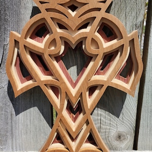May include: A layered wooden cross with intricate cut-out designs. The cross features various shades of wood, including light, medium, and dark brown, and reddish-brown. The design has a symmetrical pattern with a heart-shaped cutout in the center.