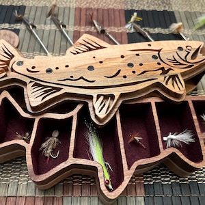 Puede incluir: Una caja de pesca con mosca de madera con forma de trucha con tapa abatible. La caja tiene seis compartimentos forrados con fieltro burdeos y contiene varios se&ntilde;uelos de pesca con mosca.