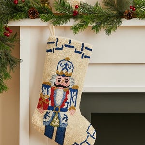May include: A cream-colored holiday stocking featuring a textured, punch-needle embroidered nutcracker design. The nutcracker wears a blue and gold uniform with a crown, holding a small red gift. Blue geometric patterns accent the top and toe.
