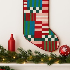 May include: A Christmas stocking with a colorful patchwork design in red, green, blue, and white. The stocking is hanging on a white wall above a garland with a red candle and ornament.