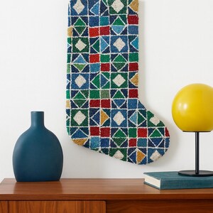 May include: A Christmas stocking with a geometric pattern in blue, green, red, yellow, and white. The stocking is hanging on a white wall. A blue vase and a yellow lamp are on a wooden cabinet below.