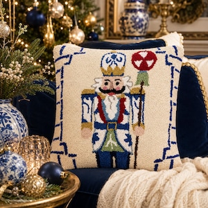 May include: A square throw pillow with a cream-colored background featuring a central hooked-yarn design of a nutcracker soldier. The soldier wears a blue, white, and red uniform with a gold crown and holds a staff topped with a red and white ornament.