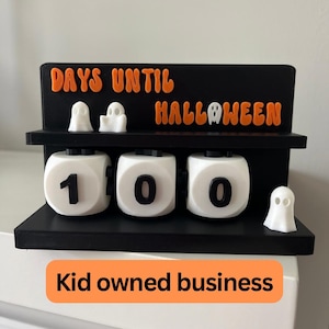 May include: A black and orange Halloween countdown calendar with the words "DAYS UNTIL HALLOWEEN". The calendar has three rotating number blocks showing "100" and small white ghost figurines. The text "Kid owned business" is at the bottom.