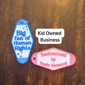 May include: Two motel-style keychains. One is blue and white with the text "Big fan of Human Rights." The other is pink and white with the text "Radicalized by basic decency." The phrase "Kid Owned Business" is also present.