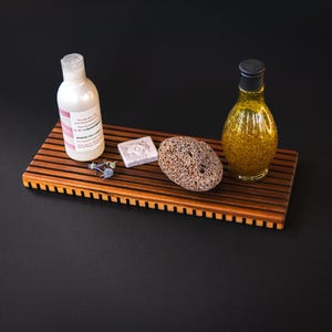 Peut inclure: Un plateau de bain en bois avec une bouteille blanche, un petit savon, une pierre ponce et une bouteille de liquide. Le plateau est en bois brun avec un design à lattes. Les articles sont disposés sur une surface noire.