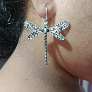 Può includere: Orecchini a forma di libellula in argento. Questi orecchini pendenti presentano un design dettagliato di libellula con intricati motivi alari e un corpo strutturato. Gli orecchini sono sospesi da un semplice gancio.