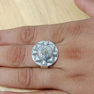 May include: A silver-toned ring featuring a large, circular face with a raised, geometric, multi-layered design. The center of the ring displays a detailed, star-like pattern surrounded by stepped, square-shaped borders, all set on a simple band.