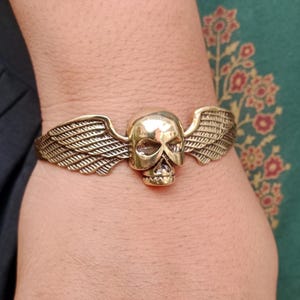Könnte beinhalten: Ein messingfarbenes Armband mit einem Totenkopf und ausgebreiteten Flügeln. Der Totenkopf weist detaillierte Gesichtszüge auf, und die Flügel sind gefiedert. Das Armband wird am Handgelenk getragen und zeigt seinen gotischen Stil und seine Handwerkskunst.