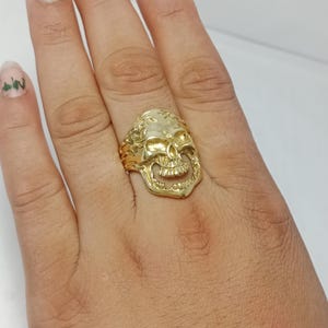 May include: A gold-toned ring featuring a detailed skull design with a polished finish. The skull occupies the center of the band, showcasing defined eye sockets, a nasal cavity, and teeth. The sides of the ring band exhibit a textured, rugged pattern.