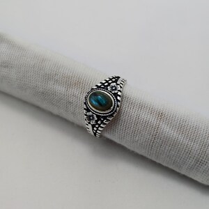 Könnte beinhalten: Ein silberner Ring mit einem ovalen, irisierenden blau-grünen Stein in der Mitte. Der Ring weist florale und geometrische Muster entlang des Bandes und um den Stein auf. Der Ring wird auf einem hellgrauen Stoff präsentiert.