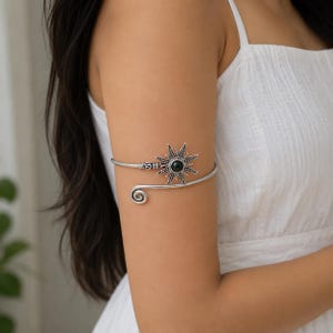 Könnte beinhalten: Ein Oberarmreif aus silberfarbenem Metall mit einem sonnenförmigen Mittelstück, das einen dunklen, runden Stein in der Mitte trägt. Das Band hat ein spiralförmiges Ende und ein texturiertes, zylindrisches Detail nahe dem Sonnenmotiv.