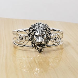 Könnte beinhalten: Ein silberfarbenes Armband mit einem zentralen Löwenkopfmotiv und detaillierter Mähne. Das Armband besteht aus dünnen, geschwungenen Metalldrähten, die auf beiden Seiten des Löwenkopfes symmetrische Spiralmuster bilden und ein offenes Design erzeugen.
