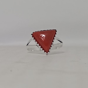 May include: A silver ring featuring a smooth, triangular-shaped red stone set in a serrated bezel. The band is polished silver and splits into two thin arms as it connects to the setting, creating a clean and simple design for this piece of jewelry.