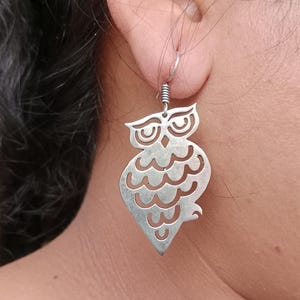 Könnte beinhalten: Silberne Eulenohrringe. Die Ohrringe haben ein ausgeschnittenes Design einer Eule mit detaillierten Augen und einem strukturierten Körper. Die Ohrringe hängen an einem silbernen Drahtbügel.