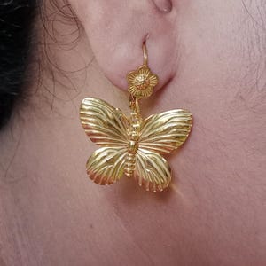 Könnte beinhalten: Ein goldfarbener Ohrring mit einem kleinen blumenförmigen Stecker an der Oberseite, der mit einem größeren, detaillierten Schmetterlingsanhänger verbunden ist. Der Schmetterling hat strukturierte, gerippte Flügel und einen segmentierten Körper.