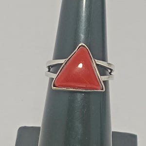 Könnte beinhalten: Ein silberner Ring mit einem auffälligen dreieckigen roten Stein. Der Ring hat ein Doppelband-Design. Der rote Stein ist in einem silbernen Band gefasst. Der Ring wird auf einem dunkelgrünen Ringhalter präsentiert, der seine kräftige Farbe und geometrische Form hervorhebt.