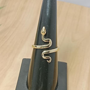 Puede incluir: Un anillo dorado con forma de serpiente, enrollado alrededor del dedo. El anillo se muestra en un soporte negro para anillos sobre una superficie de madera marrón claro. La cabeza de la serpiente está en la parte superior del anillo.