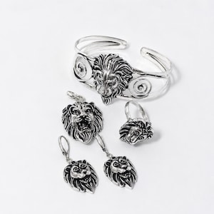 Könnte beinhalten: Ein silberfarbenes Schmuckset mit Löwenmotiv. Das Set besteht aus einem offenen Armreif mit Spiralverzierungen, einem Anhänger, einem Ring und einem Paar Ohrringen mit Klappbügel. Jedes Schmuckstück zeigt ein detailliertes, dreidimensionales Löwenkopf-Design.