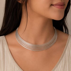 Könnte beinhalten: Eine silberfarbene Halskette im Choker-Stil, bestehend aus zehn dünnen, parallel verlaufenden Metalldrähten, die sich um den Hals legen. Die Kette hat eine polierte, reflektierende Oberfläche und wird durch kleine zylindrische Endkappen gehalten.
