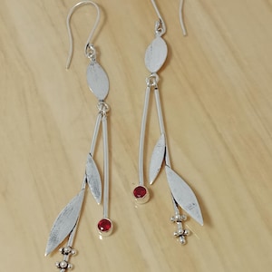 Könnte beinhalten: Silberne Hängeohrringe mit einem länglichen Design. Jeder Ohrring hat eine ovale Oberseite, einen hängenden Stiel mit einem roten Edelstein und blattförmige Akzente. Die Ohrringe haben unten eine blütenartige Ansammlung.