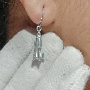 以下が含まれることがあります： 銀色のロケット型イヤリング。尖ったノーズコーン、小さな星の切り抜きがある円筒形のボディ、基部に3つの安定フィンを備えています。チャームはシンプルな湾曲した金属製のイヤーフックから吊り下げられており、光沢のある仕上げが施されています。
