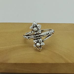 Könnte beinhalten: Ein silberfarbener Ring mit floralem Design. Der Ring hat zwei markante Blumenmotive mit detaillierten Blütenblättern und einer zentralen Perle. Das Band verläuft um den Finger, mit zusätzlichen dekorativen Elementen. Der Ring liegt auf einer Holzoberfläche.