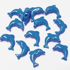 May include: A collection of glossy, blue dolphin-shaped charms. The charms are scattered across a white surface, each with a smooth, curved design. The dolphins are a consistent shade of blue, suggesting a playful or nautical theme.