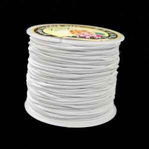 May include: A spool of white elastic cord, tightly wound and ready for use. The cord is a light gray color and appears to be made of a stretchy material. The spool has a clear plastic rim and a label with floral designs.