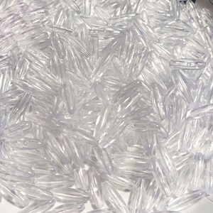 May include: A close-up shot of a pile of clear, elongated beads. Each bead has a slightly oval shape with a hole running through the center. The beads are transparent, reflecting light and creating highlights. The overall effect is a collection of small, shiny objects.