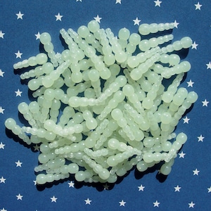 May include: A pile of pale green, glow-in-the-dark beads, each with a series of connected, rounded shapes. The beads are scattered on a blue background with white stars, creating a celestial or patriotic theme.
