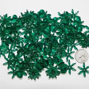 May include: A collection of translucent, emerald green flower-shaped beads. The beads are scattered across a white surface, with a US quarter coin for size comparison. The beads have a faceted design.