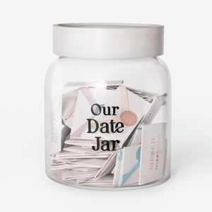 May include: A clear glass jar with a silver lid filled with folded paper notes. The words "Our Date Jar" are printed on the front. Some notes have pastel-colored accents. The jar is a decorative item.