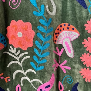May include: An olive green fabric featuring vibrant embroidery of various botanical elements and a mushroom. The design includes bright pink flowers, blue fern-like leaves, a spotted orange mushroom, and dark blue crescent shapes on a soft surface.