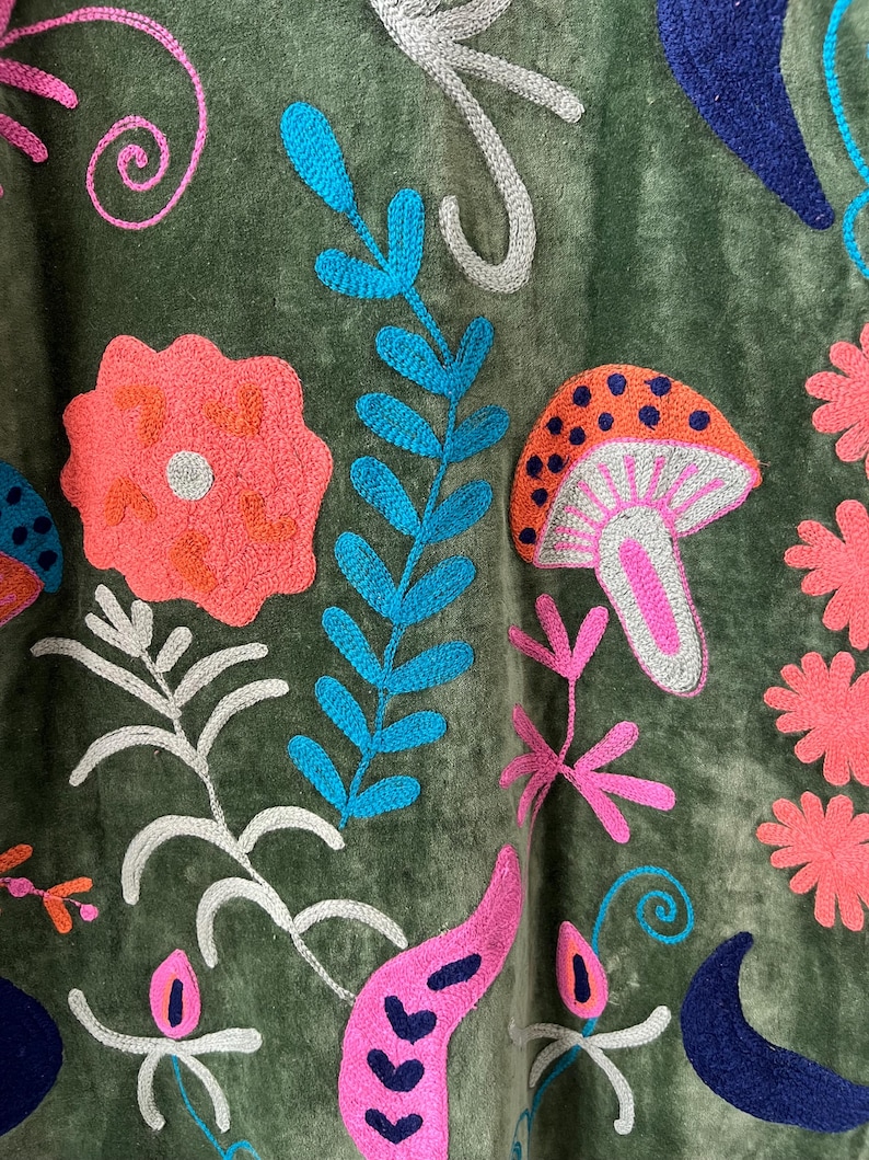 May include: An olive green fabric featuring vibrant embroidery of various botanical elements and a mushroom. The design includes bright pink flowers, blue fern-like leaves, a spotted orange mushroom, and dark blue crescent shapes on a soft surface.