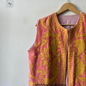 May include: A sleeveless, coral-colored vest with intricate yellow floral embroidery. The vest is reversible, with a striped lining. The garment is hanging on a wooden hanger against a white wall.