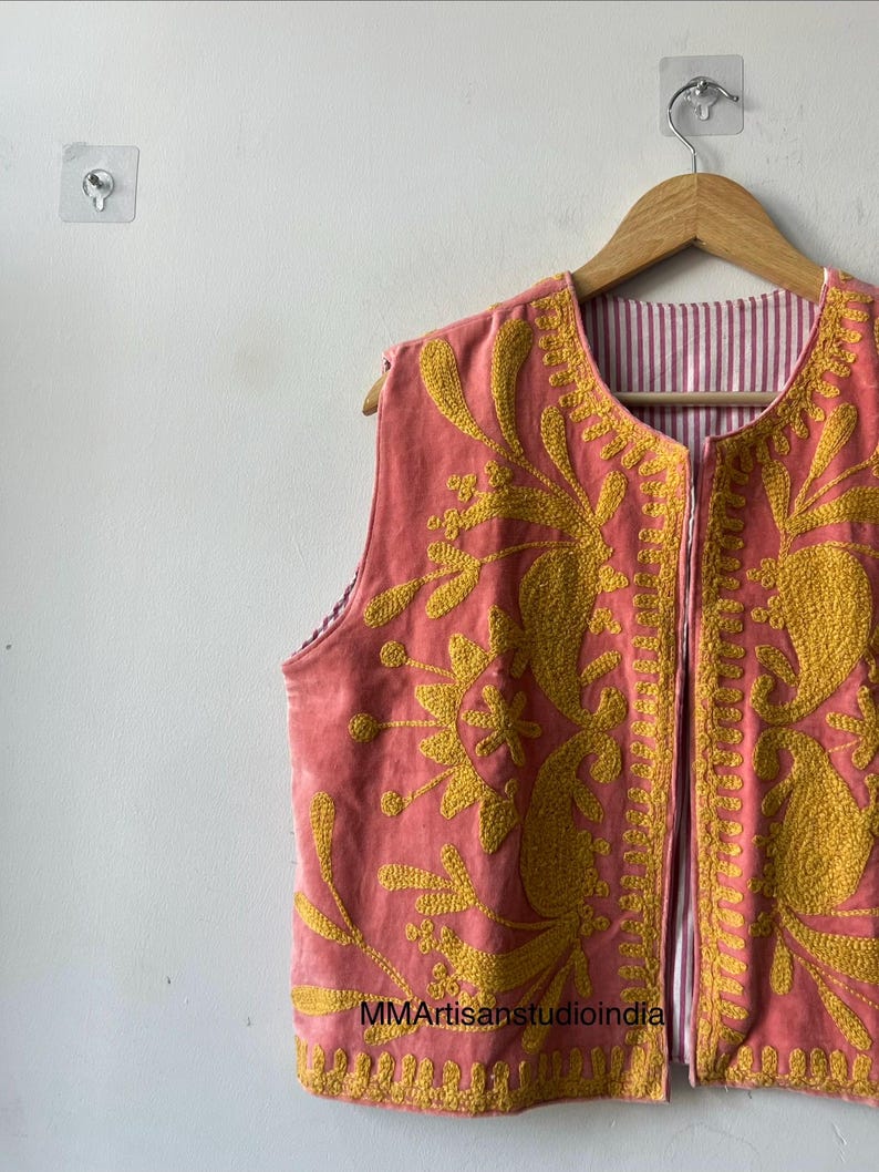May include: A sleeveless, coral-colored vest with intricate yellow floral embroidery. The vest is reversible, with a striped lining. The garment is hanging on a wooden hanger against a white wall.